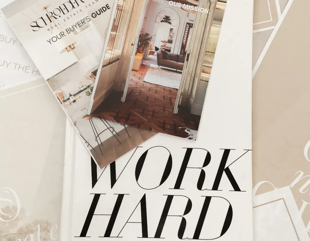 Schroeder & Co. Real Estate Team brochures, including the Buyer's Guide, resting on a white book that reads WORK HARD