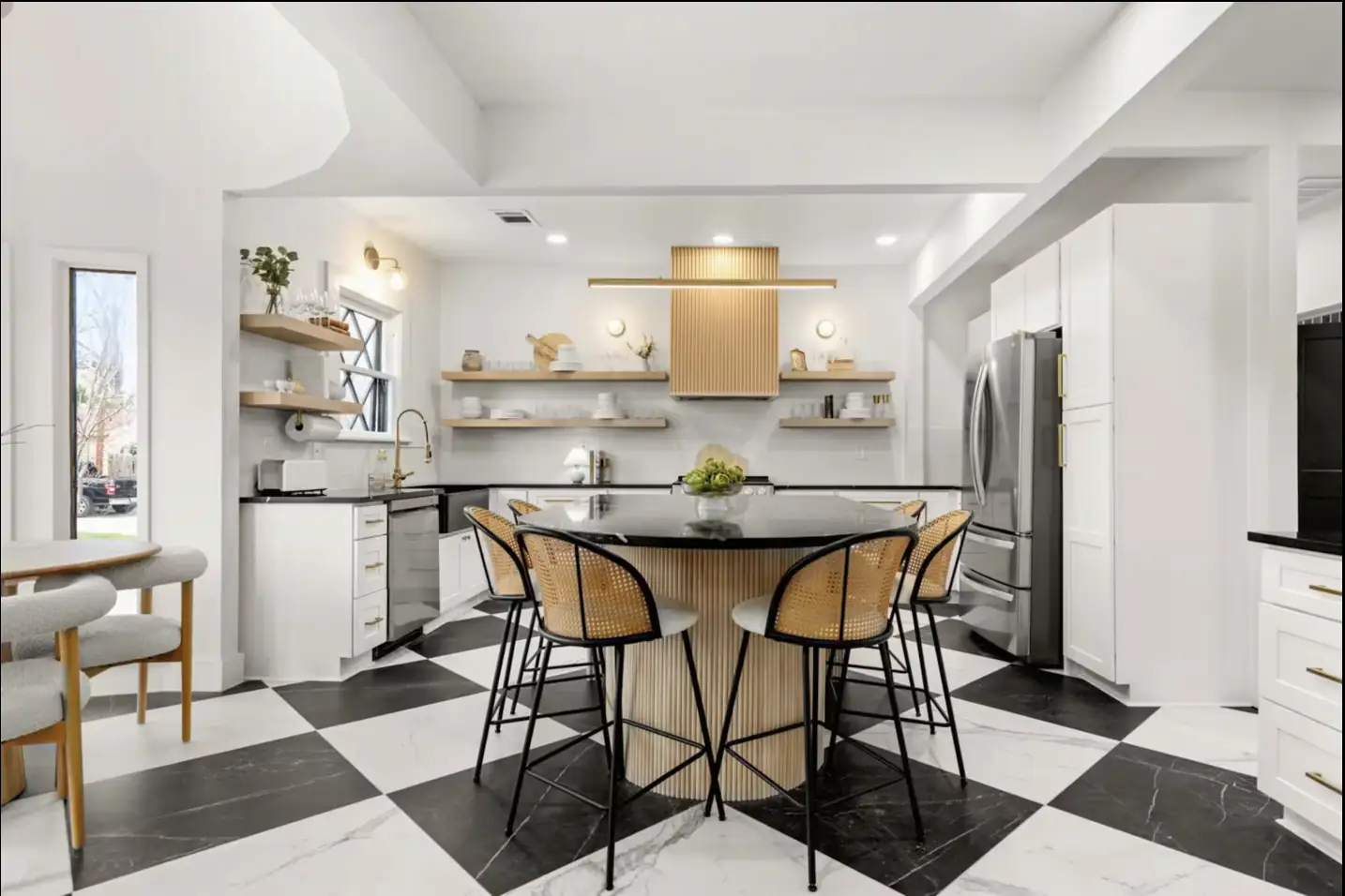 Kitchen with a black-and-white checkered floor, a round wood island with cane barstools, white cabinets with gold hardware, and open wood shelving.